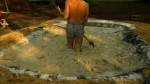 Man standing in clay, wearing short jean cut-offs with a shovel
